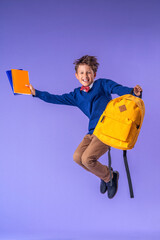 cheerful schoolboy in uniform with backpack and books jumps on purple background