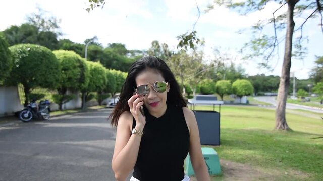 Asian woman with sunglasses walking and talking on phone. Follow shot