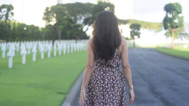 Asian woman adjusts hair and walks through cemetery. Follow shot from behind