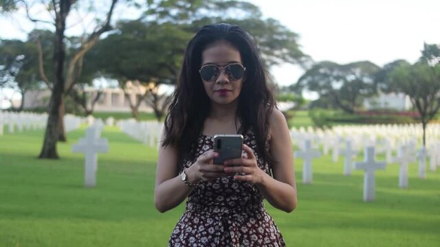 Asian woman wearing sunglasses and using cell phone in cemetery