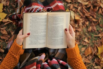  Autumn books.Reading books.Book in the hands of a girl in an orange sweater and a checkered scarf on an autumn brown foliage background. View from above.Student and learning concept.Fall season
