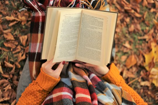  Autumn books.Reading books.Book in the hands  in an orange sweater and a checkered scarf on an autumn brown foliage background. View from above.Student and learning concept.Fall season