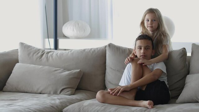 Two little cute children, sister, and brother, sitting on the sofa with pillows in the cozy room and hugging each other in front of the camera. Indoor