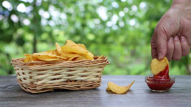 hands of adult female taking potato chips from wickered bowl and dipping them in ketchup
