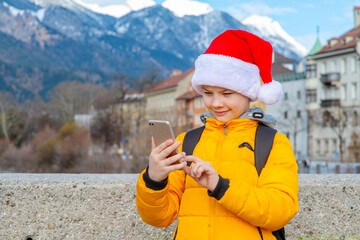 A boy in a Santa hat is looking at his mobile phone against the backdrop of the Alps in Austria. Christmas travel concept