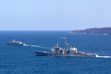 Two warships sailing out to sea past a tall coastline.