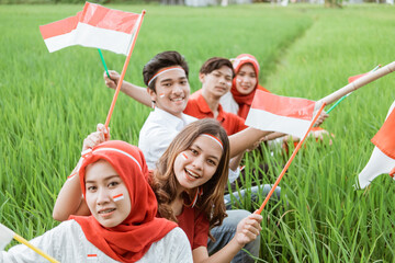 Asian youth smiles holding small red and white flags sitting in the middle of rice fields while wearing Indonesian flag attributes