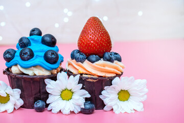 two birthday cupcakes with white daisies, decorated with blue cream blueberries and strawberries on a pink background
