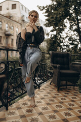 Stylish fashionable blonde woman with smoky eye makeup, in jeans, lingerie and black leather jacket on the balcony in the city. Spring autumn fashion concept. Soft selective focus.