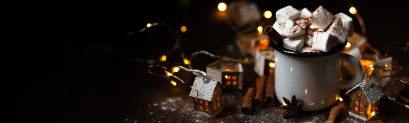 Winter hot drink: hot chocolate with marshmallows, cinnamon and anise. Christmas lights in the shape of houses as decor. Decorative snow, dark wooden background. Festive mood, cozy home atmosphere