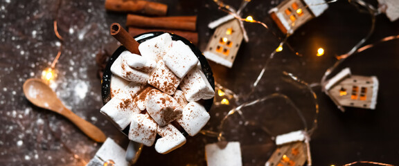 Winter hot drink: hot chocolate with marshmallows, cinnamon and anise. Christmas lights in the shape of houses. White snow, dark wooden background. Festive mood, cozy atmosphere. Flat lay top view