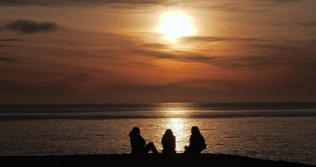 Silhouettes of three girls sitting on the beach and watching the sun rising. Friends spending time together.