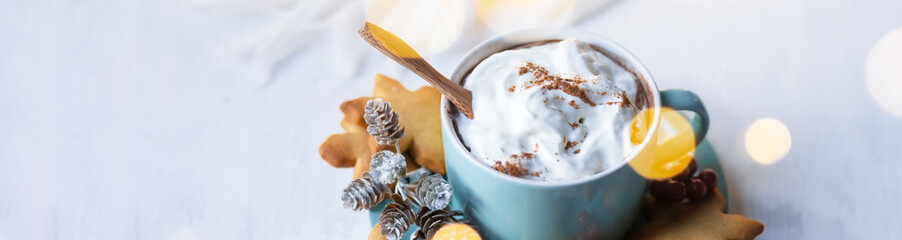 Hot winter drink: chocolate with whipped cream in blue mug. Christmas time. Cozy home atmosphere, white background. Homemade gingerbread cookies, christmas lights. Banner, copy space for text