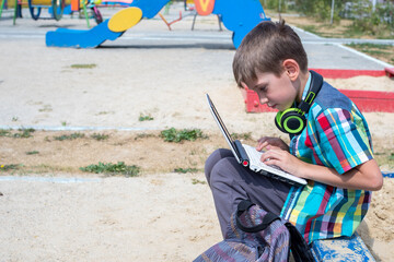 A cute boy schoolboy dressed in casual clothes is engaged on a laptop on the playground, a schoolbag lies nearby