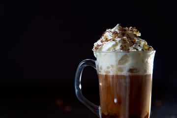Close up of a glass cup of coffee with whipped cream and chocolate on it, chocolate chip cookies and roasted coffee beans on dark background. Concept of ready to eat food, tasty snack. Selective focus
