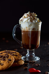 Close up of a glass cup of coffee with whipped cream and chocolate on it, chocolate chip cookies and roasted coffee beans on dark background. Concept of ready to eat food, tasty snack. Selective focus