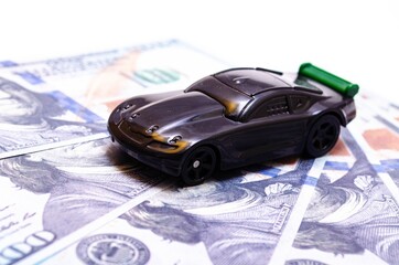 Black toy car on a heap of dollar bills on a white and black background. A sports car with a green...
