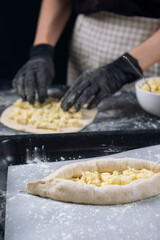 Woman in checkered apron and black gloves spread the cheese filling on the dough. Process of making bakery. Adjarian Khachapuri Recipe – Georgian cheese bread.