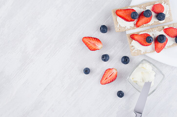 Healthy dietary breakfast of crisps rye flat toast with fresh ripe strawberry slices, blueberry, sweet cream cheese on white wood background, top view, copy space.