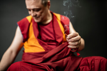Buddhist monk in red kesa, holding an incense stick with incense in his hand
