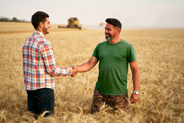 Farmer and agronomist shaking hands in wheat field after agreement. Agriculture business contract concept. Corporate farmer and landlord rancher negotiate with handshake. Combine harvesters harvest.