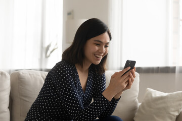 Happy millennial Asian girl sit on couch in living room texting messaging on cellphone gadget,...