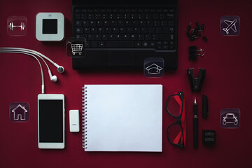 home office desk workspace with laptop, headphones, smartphone with black screen over a notebook and various icons for financial savings or spending on red background. Mockup image.