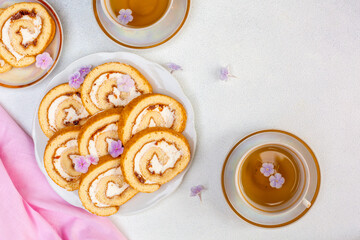 Top view off homemade roll cake with apricot marmalaid and cheese cream and cups with tea. Decorated with pink flowers. Copy space.