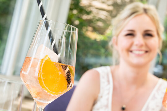 Apperitiv mit Orangenscheibe im Vordergrund, l&auml;chelnde junge blonde Frau im Hintergrund