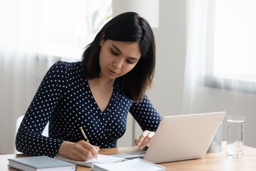 Concentrated Asian millennial girl sit at desk study make notes using modern laptop, focused Vietnamese young woman handwrite take online course or distant training on internet on computer from home
