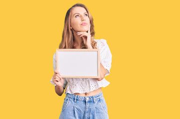 Fototapeta na wymiar Young caucasian woman with blond hair holding blank empty banner serious face thinking about question with hand on chin, thoughtful about confusing idea
