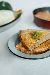 piece of vegan vegetable pie with cheese and greens on a blue ceramic plate on white cement, zucchini and green pumpkin, the shape with the whole pie is blurred in background, vertical, vegetable menu