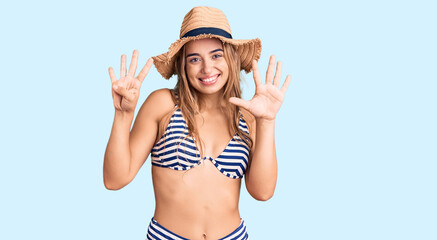 Young beautiful blonde woman wearing bikini and hat showing and pointing up with fingers number nine while smiling confident and happy.