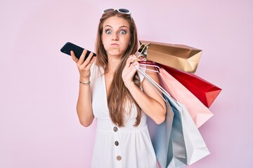 Young blonde girl holding shopping bags talking on the smartphone puffing cheeks with funny face. mouth inflated with air, catching air.