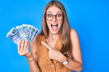 Young blonde girl holding south african rand banknotes celebrating crazy and amazed for success with open eyes screaming excited.
