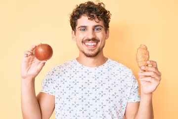 Young caucasian man with curly hair holding apple and croissant smiling with a happy and cool smile on face. showing teeth.