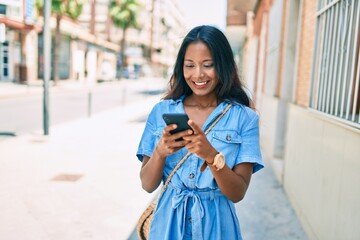 Fototapeta premium Young beautiful indian woman smiling happy using smartphone walking at the city