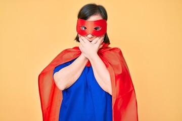 Brunette woman with down syndrome wearing super hero costume shocked covering mouth with hands for mistake. secret concept.