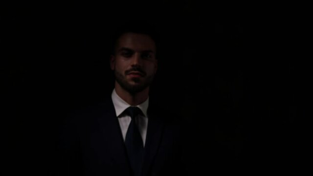sexy young businessman coming from the dark, exposing himself, fixing suit and tie, getting fingers through hair, holding hands to face and touching lips and beard, walking and fading away