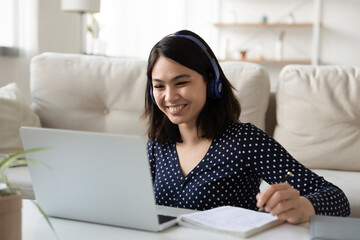 Happy millennial Asian girl in headphone use laptop take online course training make notes, smiling young Vietnamese female student in earphones study on computer from home, distant education concept