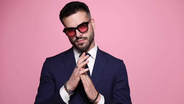 young handsome businessman with sunglasses is holding his hands together, slowly rubbing his fingers and looking at camera with a seductive look, pulling an invisible rope on pink background