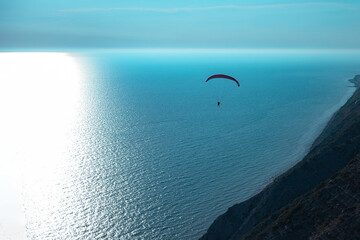 Silhouette of a flying paraglider on a background of blue sea near the coast. The black silhouette of a paraglider flies near the seashore.