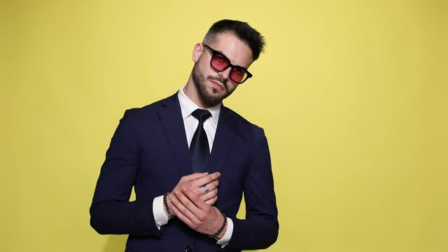 sexy unshaved businessman in suit wearing sunglasses touching hands and rubbing palms,looking to side and down on yellow background