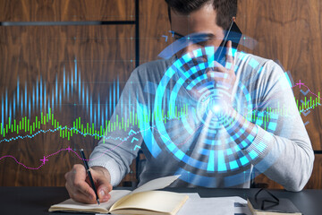 Handsome analyst in casual long sleeve talking phone, taking notes at office workplace try to find the best solution for client capitalisation. Double exposure. Financial chart hologram.