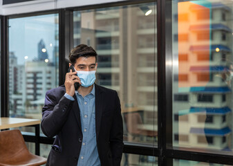 Young CEO or businessman with surgical face mask using mobile phone during office hours as part of Business New Normal and lifestyle during COVID-19 or Coronavirus outbreak. Man wearing face mask