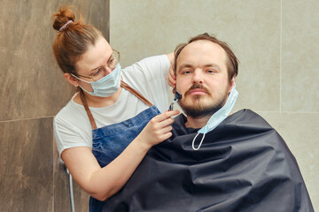 Obraz premium Woman in medical mask shaves man in home bathroom with safety razor. Concept of self-cutting and shaving during the flu epidemic