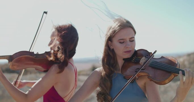 Close view of female violinists duet in blowing dresses play among cliffs