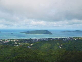 View to the sea coast, water, horizon and green island from the top. Panorama of ocean and cloudy sky. Rain weather. Green trees, forest. Coastal town on the shore. Rainforest, jungle. Paradise