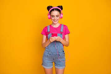 Photo of cheerful lady hold telephone listen music modern technology earphones choose song to turn on wear pink t-shirt denim overall isolated yellow bright color background