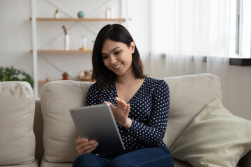 Smiling Asian millennial girl sit relax on couch in living room browse internet on modern tablet gadget, happy Vietnamese young woman rest on sofa at home using pad device, technology concept